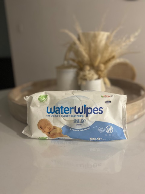A pack of WaterWipes, labeled as 'The World's Purest Baby Wipes,' featuring a baby graphic and highlighting that they contain 99.9% water and a drop of fruit extract. The background shows a modern, softly lit setting with decorative elements.