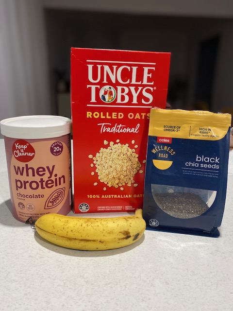 A collection of ingredients for a protein-rich breakfast: a container of chocolate whey protein, a box of Uncle Toby's rolled oats, a bag of black chia seeds, and a ripe banana, all displayed on a kitchen countertop.