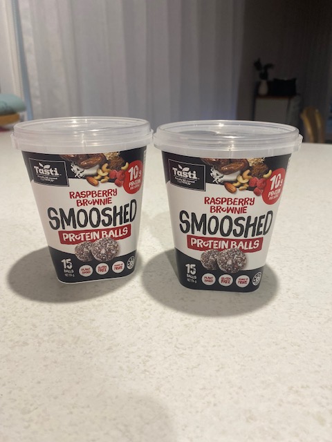 Two containers of Tasti Smooshed Raspberry Brownie Protein Balls sitting on a light-colored countertop.