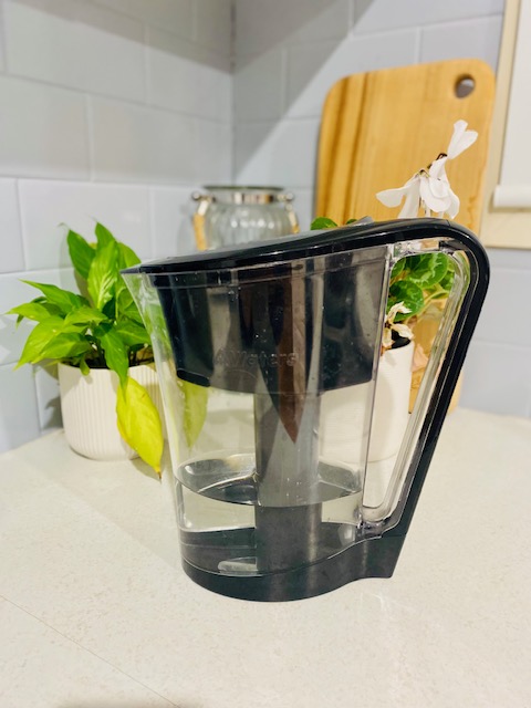 A Waters Co water filter jug sits on a kitchen countertop next to green plants and a wooden cutting board.